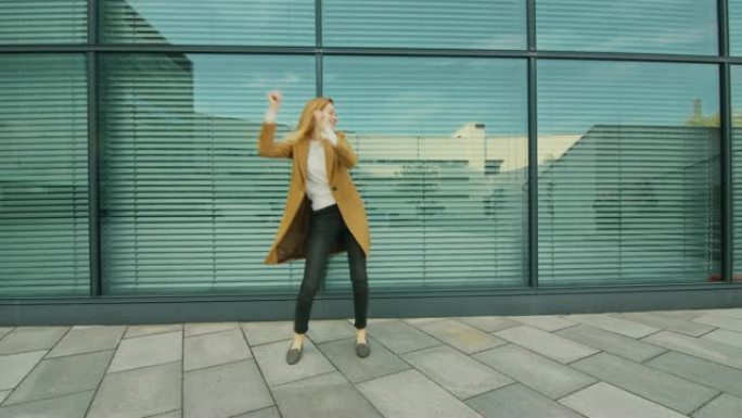 开朗快乐的年轻女子在玻璃建筑前积极跳舞。她穿着一件棕色长外套。在商务中心旁边拍摄的场景。天很亮。