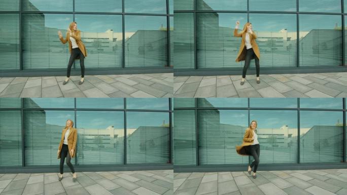 开朗快乐的年轻女子在玻璃建筑前积极跳舞。她穿着一件棕色长外套。在商务中心旁边拍摄的场景。天很亮。