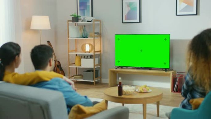 在家里，一群不同的朋友在绿色色度键屏幕电视上观看喜剧，坐在客厅里吃零食和喝饮料。年轻人玩得开心。