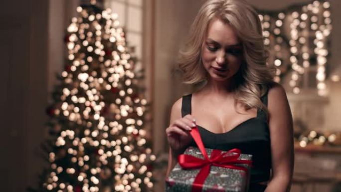 一个穿着晚礼服的美丽金发女郎在圣诞树的背景上看着她的礼物。