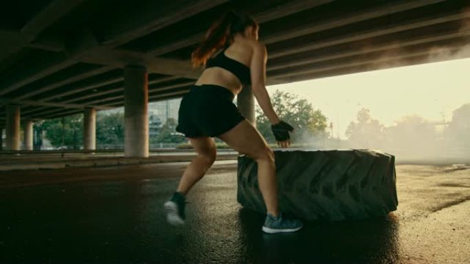 穿着黑色运动上衣和短裤的美丽健身女孩正在街上做运动。她在城市环境中翻了个沉重的大轮胎