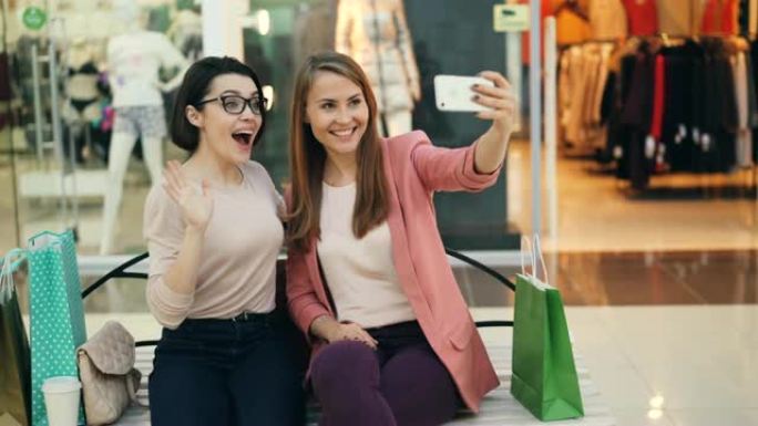 开朗的女性朋友正坐在购物中心的长凳上，用智能手机自拍，并戴着眼镜摆姿势，摆出有趣的面孔。照片、商店和