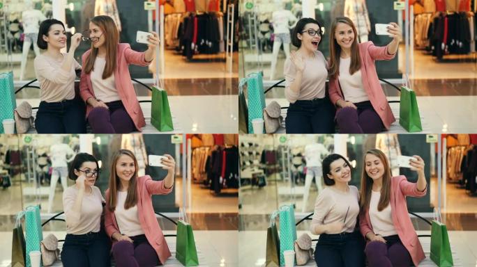 开朗的女性朋友正坐在购物中心的长凳上，用智能手机自拍，并戴着眼镜摆姿势，摆出有趣的面孔。照片、商店和