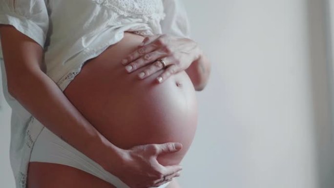 一位穿着白色连衣裙的年轻孕妇的真实特写镜头正在抚摸她的肚子，她早上刚在卧室醒来