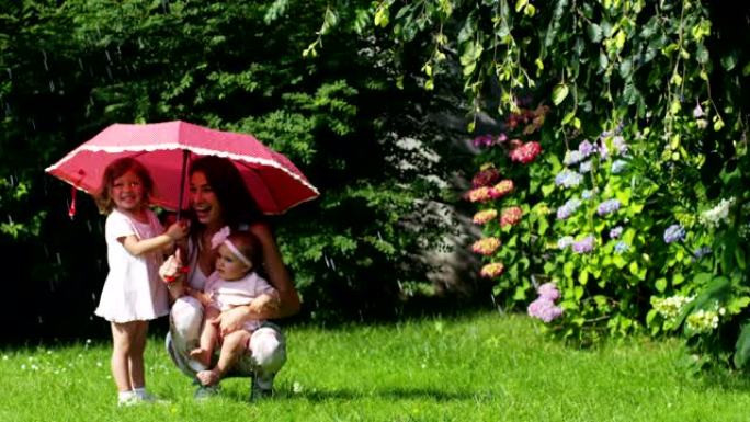 花园里的小女孩和妈妈一起躲在红色的雨伞下玩耍，对着溅水，看着镜头。