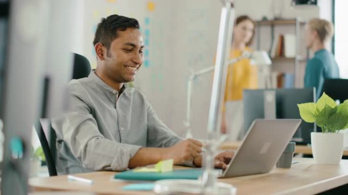 坐在办公桌前的聪明英俊的印度上班族在笔记本电脑上工作。成功的人会做 “是” 的手势并庆祝。在现代办公