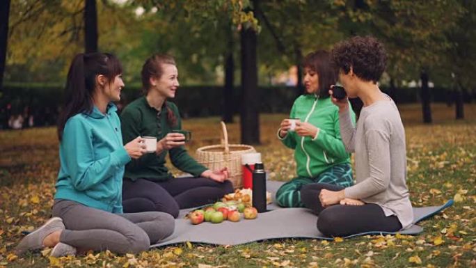 快乐的女孩在公园里野餐，坐在瑜伽垫上，在秋天的户外练习后吃饭，女孩们在说笑。交流和食物概念。