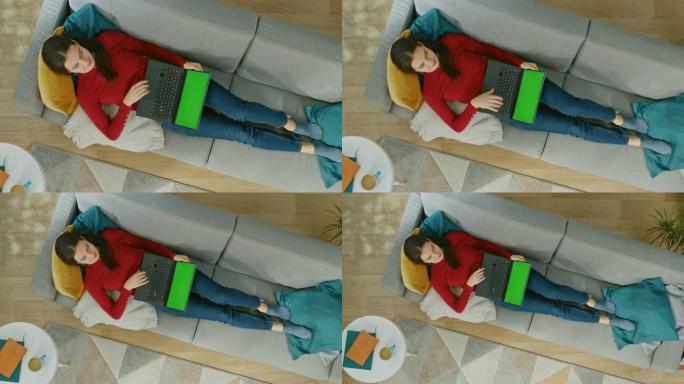穿着红色套头衫和蓝色牛仔裤的快乐年轻女孩躺在沙发上，使用带有绿色屏幕的笔记本电脑。舒适的客厅，配有现