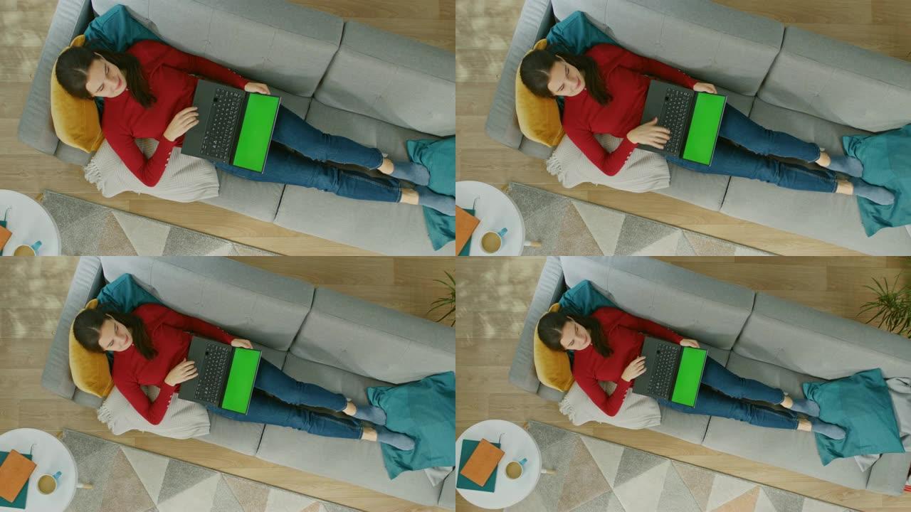 穿着红色套头衫和蓝色牛仔裤的快乐年轻女孩躺在沙发上，使用带有绿色屏幕的笔记本电脑。舒适的客厅，配有现