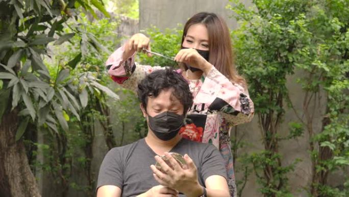 年轻的亚洲妇女在保护性医疗面罩做理发与剪刀她的男友因为封锁。居家隔离冠状病毒大流行预防，社会隔离概念