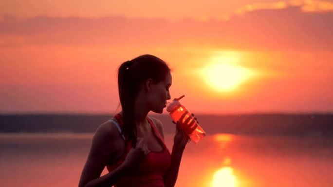 马尾辫的漂亮女士站在湖边，喝瓶子里的淡水。