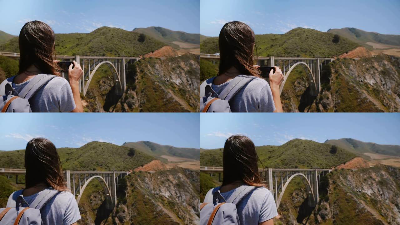 背景带背包的年轻旅游女子在标志性的比克斯比峡谷大桥大苏尔拍摄史诗般的风景的智能手机照片