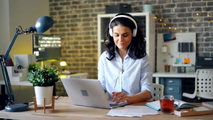 戴着耳机的迷人女士在工作中听音乐和使用笔记本电脑