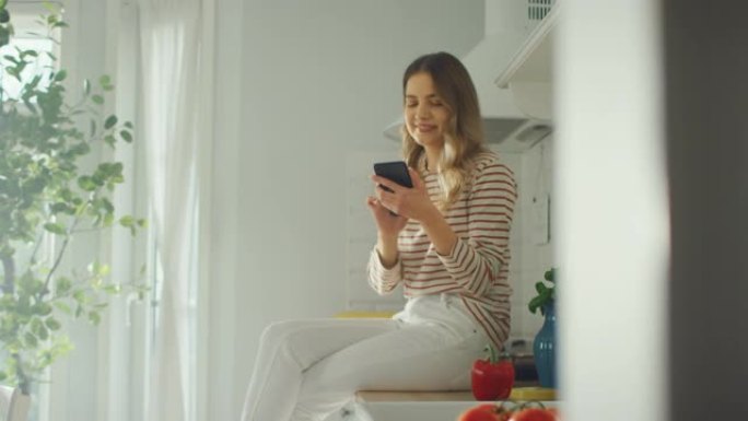穿着条纹套头衫和白色裤子的美丽年轻女性正坐在厨房家具上，用她的智能手机在一个现代阳光明媚的厨房里。她