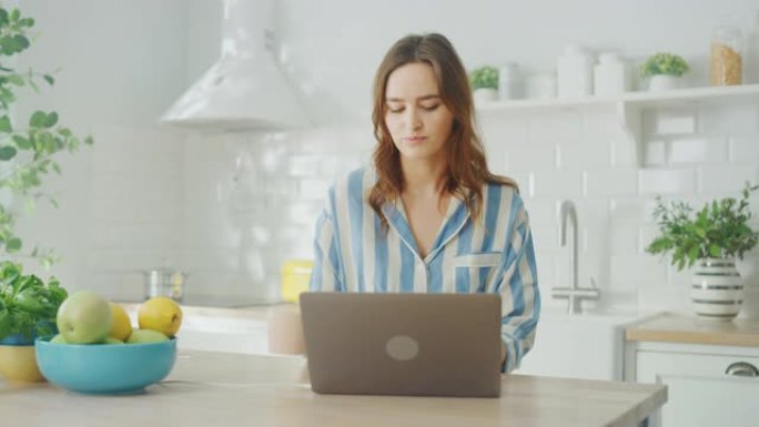 年轻漂亮的女人使用笔记本电脑，早上喝杯咖啡或茶，同时穿着蓝色睡衣。黑发女性坐在现代阳光明媚的厨房里。