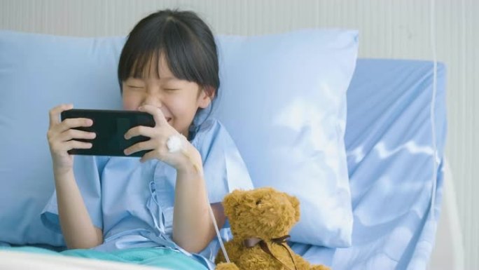 可爱的小女孩躺在医院的床上，看有趣的卡通，智能手机上的电影。疾病和治疗。