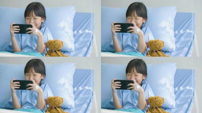 可爱的小女孩躺在医院的床上，看有趣的卡通，智能手机上的电影。疾病和治疗。
