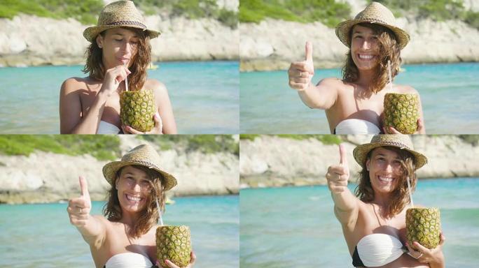 美丽的年轻女孩在海边喝菠萝鸡尾酒，穿着白色帽子的泳衣，金色的沙子，大海的背景。