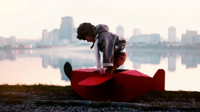 小快乐飞行员男孩在惊人的宁静城市湖全景慢动作中穿上有趣的红色纸板飞机服装。