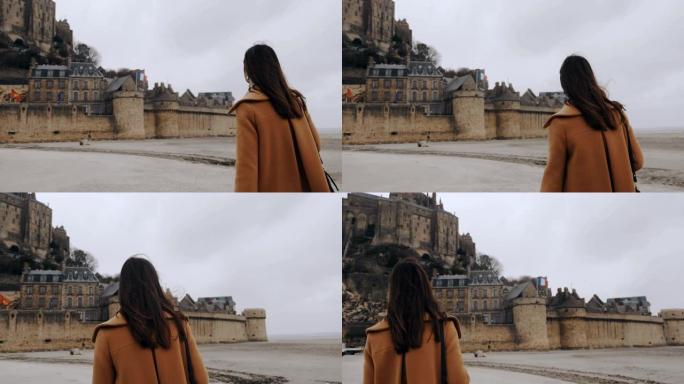 镜头跟随美丽快乐的旅游女人在阴天慢动作中走到史诗般的圣米歇尔山城堡附近。
