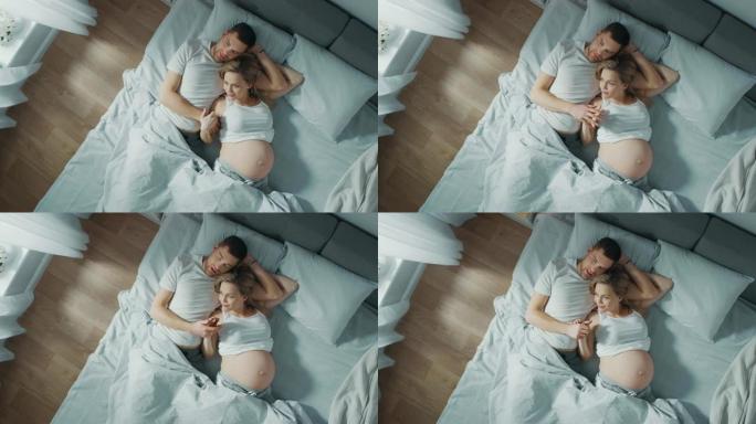幸福的年轻夫妇在床上拥抱在一起，年轻的女人怀孕了，有爱心的伴侣温柔地抚摸着她的肚子。自上而下缩小相机