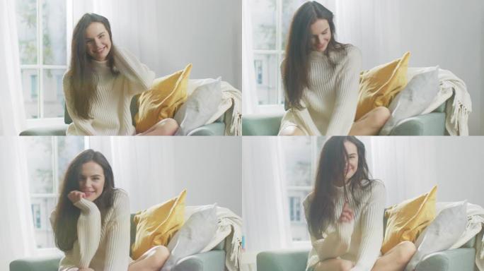 美丽的年轻女子坐在椅子上对着镜头迷人地微笑。周日早上，穿着超大号白色针织毛衣的可爱的女孩在舒适的公寓