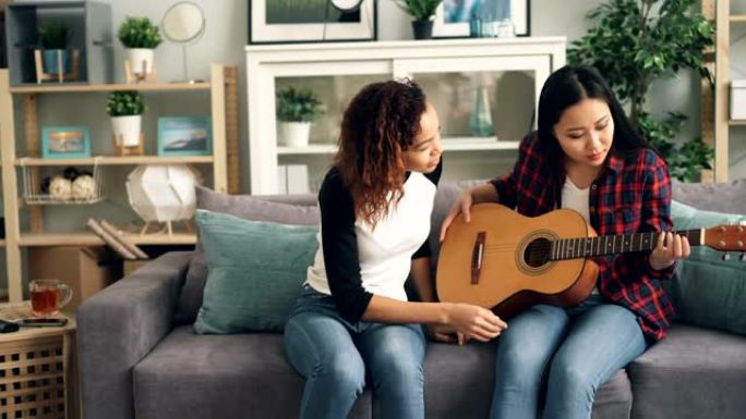 开朗的非裔美国女孩正在教她的亚洲朋友在家弹吉他。年轻妇女坐在沙发上拿着乐器说话。