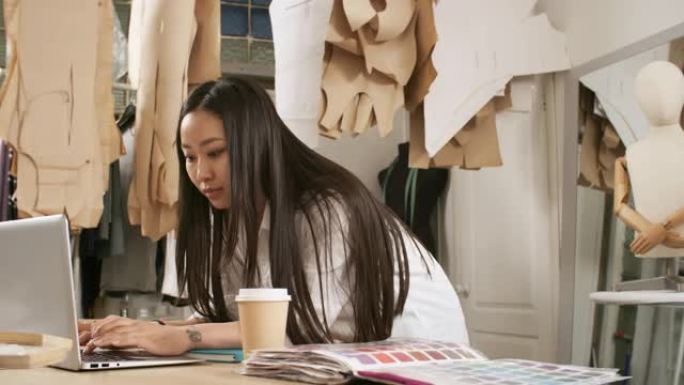 亚洲女裁缝将在缝纫工作室工作，并在笔记本电脑上打字
