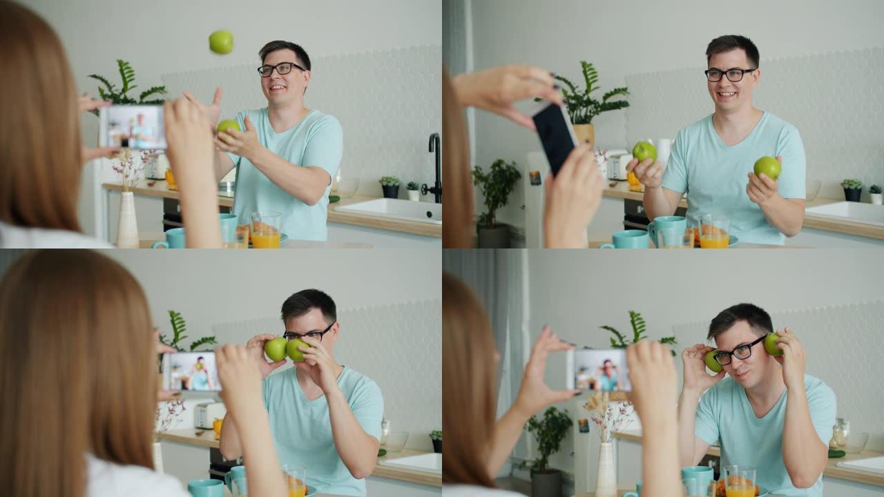 快乐的男人在厨房里玩水果，而妻子用智能手机拍照