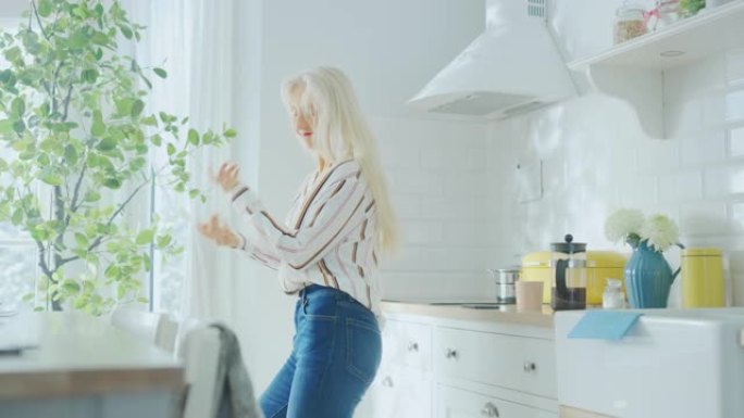 穿着蓝色牛仔裤和白色条纹衬衫的正宗高级女性正在厨房里创造性地跳舞。阳光现代厨房，拥有健康的生活方式氛