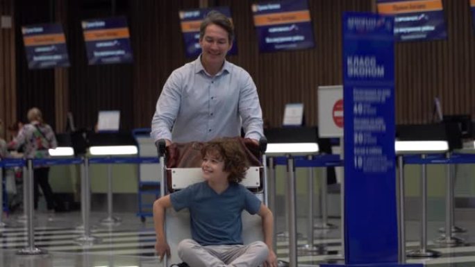 儿子坐在机场时，顽皮的爸爸推着行李车既兴奋又快乐