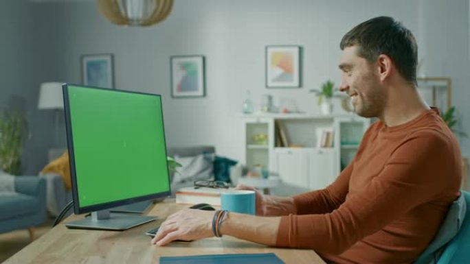 坐在家里办公桌前的英俊男人使用带有绿屏模型的个人计算机。他喝杯子里的饮料。