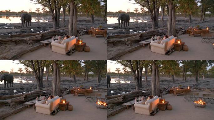 4k视角的游客坐在日落时分，傍晚的大火点燃，看着大象在博茨瓦纳Khwai私人保护区前的水坑里喝酒