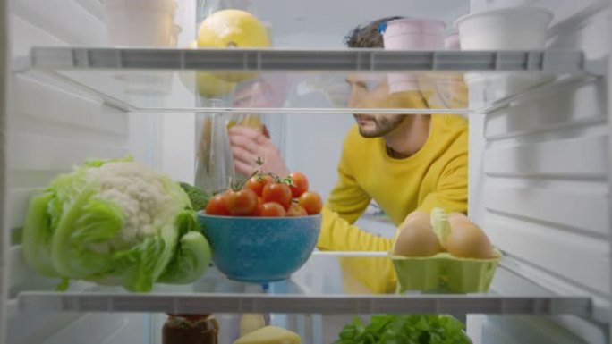 厨房冰箱内的摄像头: 男人打开冰箱门，看着里面拿出水瓶。保持水分的健康人。从装满健康食品、食品杂货、