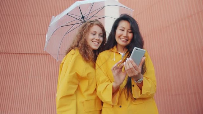 穿着雨衣的开朗年轻女性在户外伞下使用智能手机