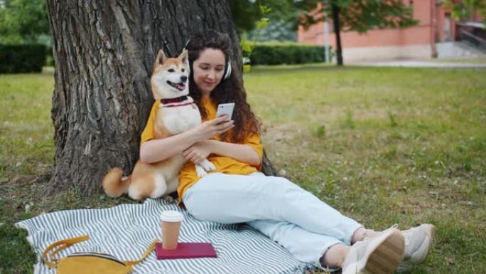 现代女孩在公园里用智能手机带着狗用耳机听音乐