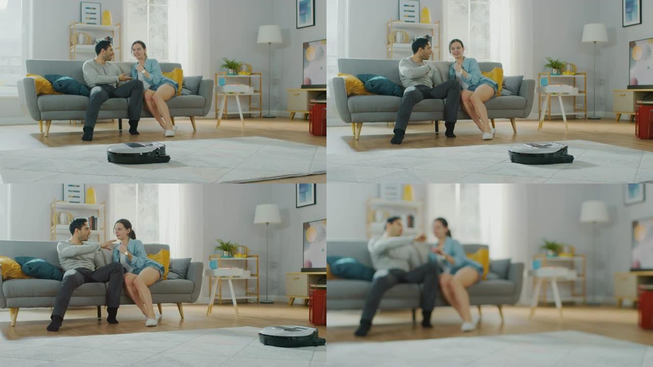 智能机器人真空吸尘器吸走地毯上的灰尘。美丽的夫妇坐在沙发上，在后台聊天。科技家电设备超越了它们。