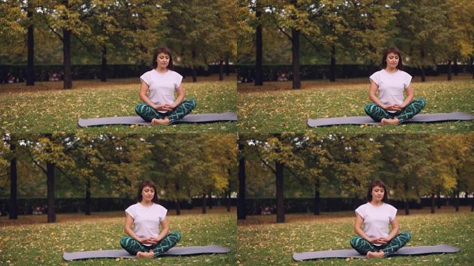 漂亮的年轻女子正在公园坐在瑜伽垫上的莲花姿势放松，并在单独练习后呼吸休息。冥想和自然概念。