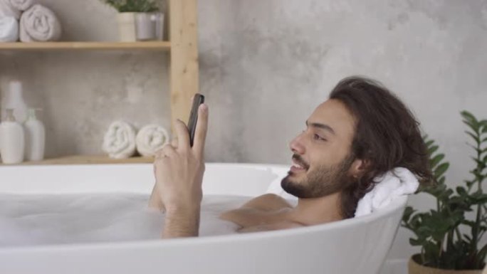 躺在浴缸里使用智能手机的快乐男人