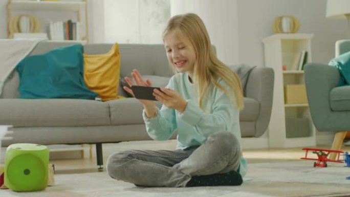 聪明可爱的女孩坐在地毯上玩智能手机上的视频游戏，在水平景观模式下拿着和使用手机。孩子在阳光明媚的客厅