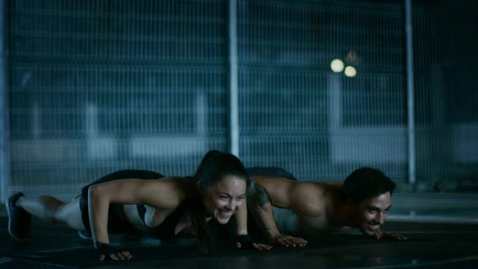 微笑快乐的运动健身夫妇做俯卧撑练习并击掌。锻炼是在有围栏的室外篮球场进行的。居民区雨后的夜间录像。