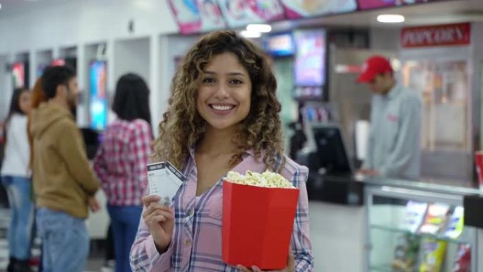 美丽的拉丁美洲年轻女子在电影院拿着爆米花和门票面对镜头微笑