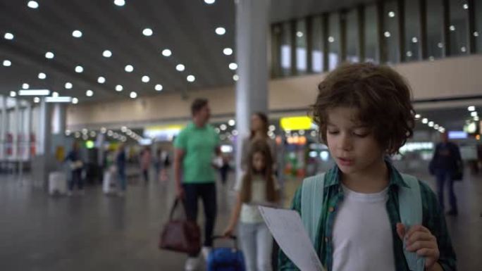 可爱的小男孩在他身后的机场和家人散步时看着他的登机牌