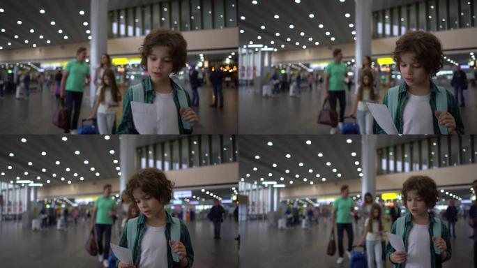 可爱的小男孩在他身后的机场和家人散步时看着他的登机牌