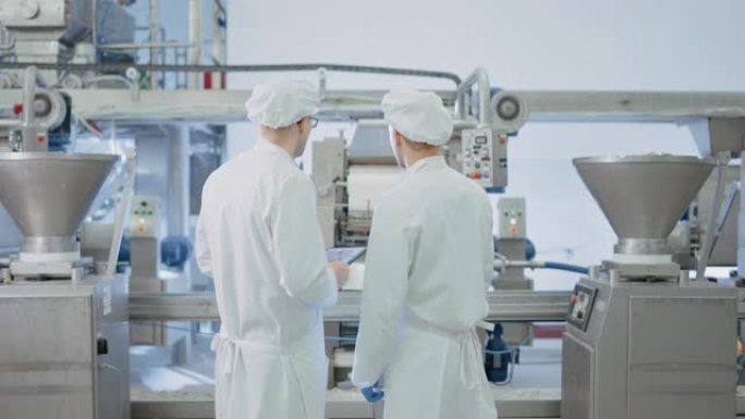 两名年轻的食品厂员工讨论工作相关事宜。男技术员或质量经理使用平板电脑工作。他们戴着白色的卫生帽子和工