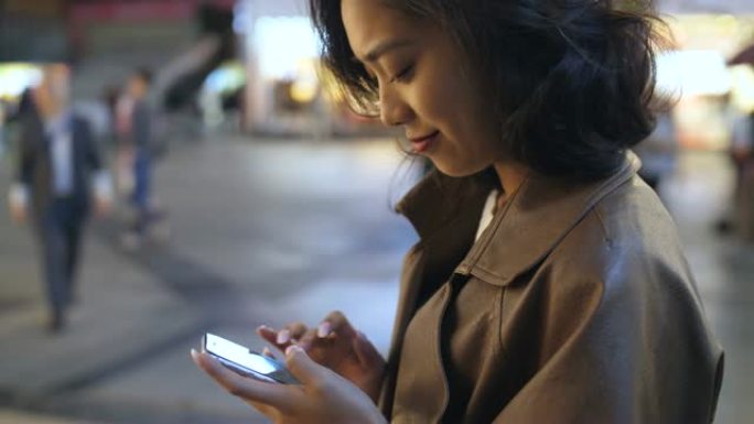 漂亮年轻的亚洲女人晚上在城市街道上使用手机，4k