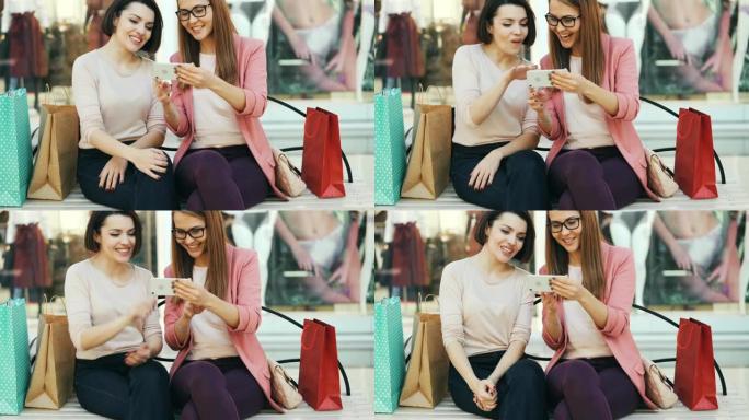 情绪化的女孩看着智能手机屏幕聊天，笑着坐在购物中心的长凳上，带着包，玩得开心。现代技术和通信概念。
