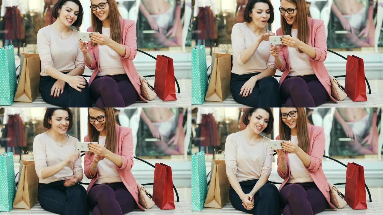 情绪化的女孩看着智能手机屏幕聊天，笑着坐在购物中心的长凳上，带着包，玩得开心。现代技术和通信概念。