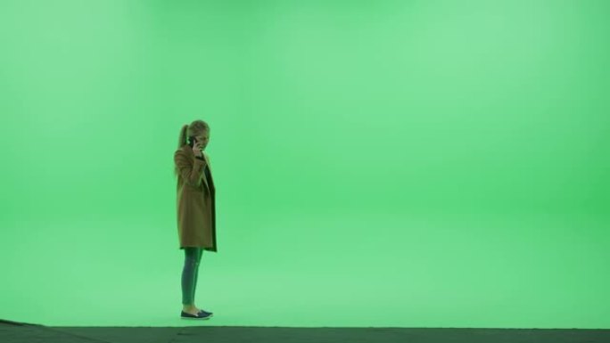 绿屏色键工作室: 漂亮的年轻女子穿着时尚的春装，使用智能手机，拿着手机聊天穿过房间。侧视图摄像机拍摄