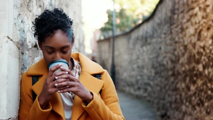 千禧一代黑人妇女穿着黄色外套，靠在小巷里的石墙上喝外卖咖啡，专注于前景